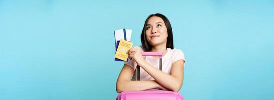 Happy asian female tourist dreaming of trip, lean on suitcase, holding covid-19 international vaccination certificate, two tickets and passport, thinking of journey, blue background photo