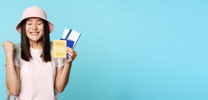 Happy asian girl triumphing, showing vaccination certificate, health passport and flight tickets, celebrating about trip, tourist feeling joy, standing over blue background photo