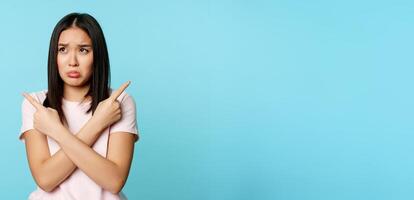 Confused and sad asian girl pointing sideways, looking clueless or indecisive, problem with choosing, pulling long upset face, blue background photo