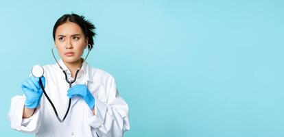 Image of serious medical worker, asian doctor listening to patient lungs with stethoscope, examining person, standing over blue background photo