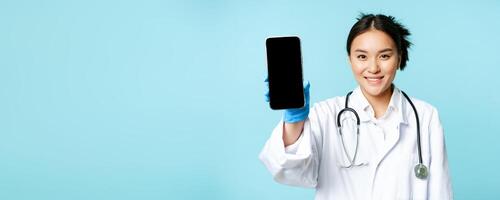 Web healthcare, online help concept. Smiling asian woman doctor, showing smartphone app interface, mobile screen, standing in uniform over blue background photo