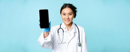 Web healthcare, online help concept. Smiling asian woman doctor, showing smartphone app interface, mobile screen, standing in uniform over blue background photo