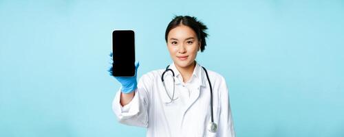 Web medical doctor concept. Smiling asian physician showing mobile phone screen, standing in clinic uniform, blue background photo