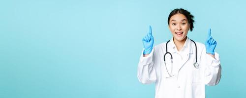 Enthusiastic asian doctor, female physician pointing fingers up and smiling, showing advertisement, demonstrating promo, blue background photo