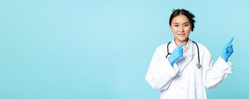 Smiling asian healthcare worker, wearing sterile gloves and uniform, pointing right, demonstrating chart, hospital advertisement, blue background photo