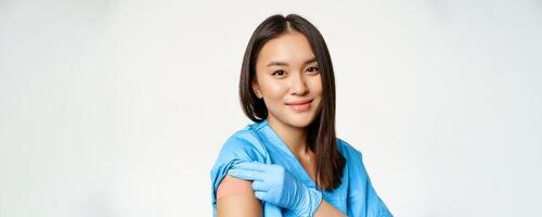 Vaccination program and covid-19 concept. Portrait of asian healthcare worker in medical robe, showing vaccinated arm and smiling, standing over white background photo