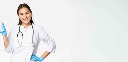 Smiling asian physician holding hand sanitizer in rubber gloves, showing bottle with antiseptic for coronavirus prevention, white background photo