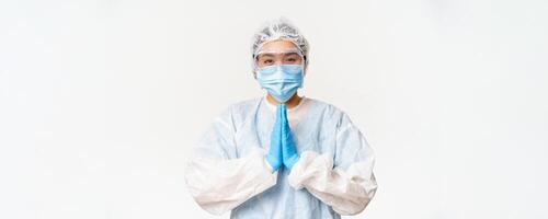 Please vaccinate. Smiling asian female doctor or nurse in personal protective equipment, showing beg, asking gesture, standing over white background photo