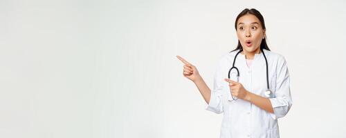 Portrait of enthusiastic female doctor, asian physician pointing and looking left with surprised, amazed face expression, standing in medical robe against white background photo