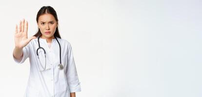 Asian doctor shows palm, stop gesture. Serious displeased female physician extend her arm, prohibit action, taboo sign, standing in uniform against white background photo