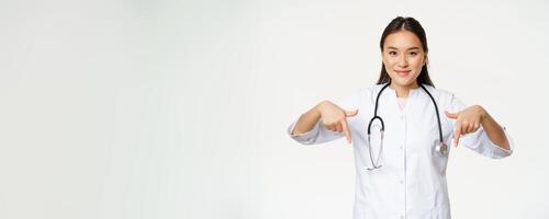 Young asian female doctor, pointing fingers down and smiling, wearing stethoscope and medical uniform, standing over white background photo