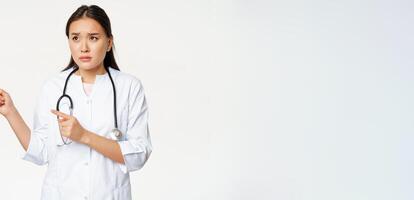 Skeptical female doctor, concerned nurse pointing and looking left with worried face expression, standing in white medical robe against studio background photo
