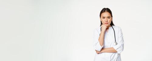 Image of angry nurse, asian female doctor looking annoyed and bothered, furrow eyebrows and sulking thoughtful, standing over white background photo