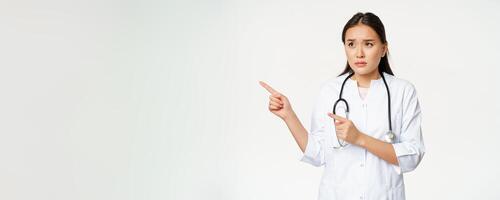 Skeptical female doctor, concerned nurse pointing and looking left with worried face expression, standing in white medical robe against studio background photo