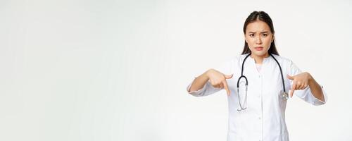 Disappointed asian female doctor pointing fingers down, frowning and looking concerned, showing bad product, standing over white background photo