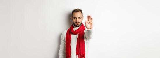 Serious bearded guy saying no, showing stop sign, rejection sign, prohibit action, standing in winter sweater and red scarf against white background photo