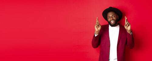 Hopeful african american man making wish, holding fingers crossed for good luck and smiling optimistic, standing against red party background photo