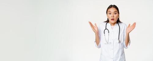 Image of surprised doctor, raising hands up and looking shocked, standing in medical hospital uniform, white background photo