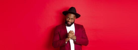 Cunning black man having an idea, smiling devious and rubbing hands, standing against red background and scheming photo
