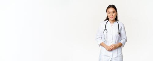 Smiling asian medical worker with stethoscope, wearing doctor uniform, looking helpful at patient, standing over white background photo