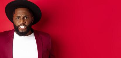 Tensed african american man clench teeth and frowning, looking at something uncomfortable and cringe, standing over red background photo
