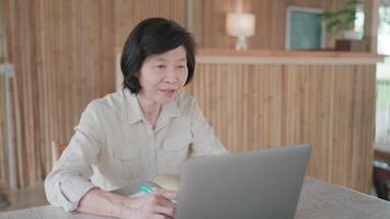 Close up of senior woman face using pen to take notes and using laptop to talk on call on table in bamboo house. Researching, senior citizen learning to cook online. Self development concept. video