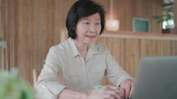 Mother is having call on laptop using pen to take notes and there is a book on the table in bamboo house, learning and researching online, call with family, technology and internet video