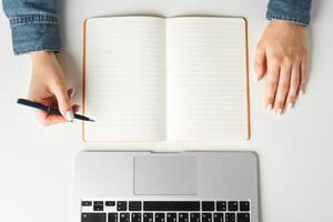 Female hand writing in a notebook on table with laptop, view from above photo