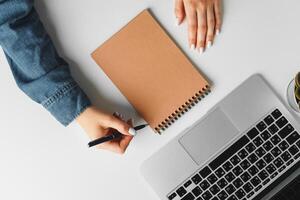 Female hand writing in a notebook on table with laptop, view from above photo