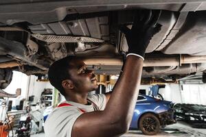 Young African mechanic in uniform working under the car in car service center photo