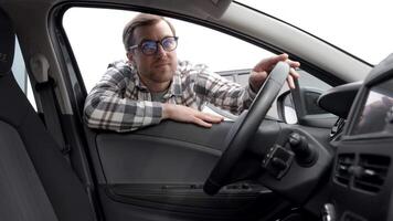 Young man looking through window inside new car, checking it before purchase at modern dealership store. Cheerful guy buying or renting automobile at showroom video