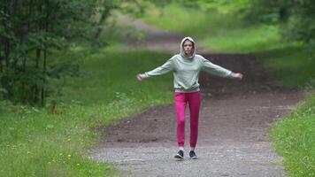 Athletic woman dressed in sports clothing - hoodie, joggers and sports shoe walking on pathway in park and doing warm-up exercises during summer fitness training. Full length view, real time video