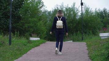 Rear view of an unrecognizable woman walking along pathway in park in summer. Model is wearing blue jacket, jeans and sneakers, is carrying small backpack on her back. Handheld, real time, zoom in video