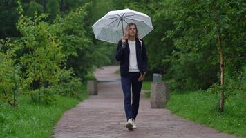 Full length front view of woman walking on sidewalk in park with transparent umbrella in her hand in rainy summer weather. Candid model in blue jacket, jeans and pullover. Handheld shot, zoom out video
