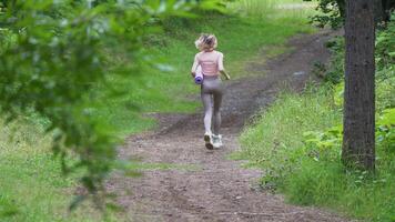 Full length view of female fitness model running along pathway in summer park during sports training, moving activity. Rear view of unrecognizable runner dressed in sportswear and carries exercise mat video