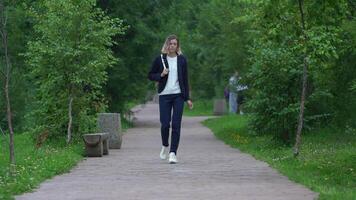 Full length view of loneliness woman walking on footpath in public park in summertime. Beautiful model dressed in blue jacket and jeans and is carrying small backpack on back. Handheld shot, zoom out video