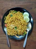 Flat lay view of delicious fried noodles with some slices of fresh cucumber on a black ceramic plate. photo