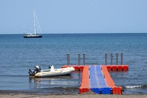 A red and blue pontoon pier on the beach a boat and yacht in the background. A floating plastic dock on a beautiful bay. photo
