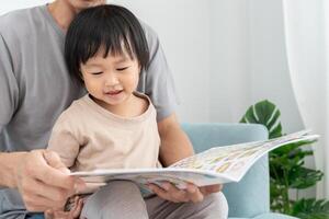 father relax and read book with baby time together at home. parent sit on sofa with daughter and reading a story. learn development, childcare, laughing, education, storytelling, practice. photo
