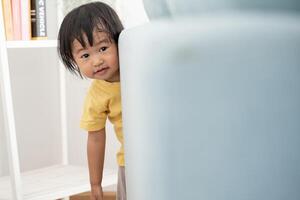 little asian children kid playing hide and seek standing behind the sofa, Adorable child having fun in the home. happy family have fun with kids, activity, learning, activity, game, meditation photo