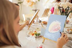 Close-up of canvas on easel and hand with spatula. Young artist paints picture at table. Blonde in painting workshop. Girl is learning to draw in art studio. photo