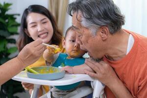 un pequeño niño comiendo su primero comida, padres y abuelos animar emocionado foto