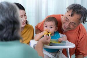 un pequeño niño comiendo su primero comida, padres y abuelos animar emocionado foto