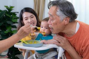 un pequeño niño comiendo su primero comida, padres y abuelos animar emocionado foto
