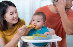 un pequeño niño comiendo su primero comida, padres y abuelos animar emocionado foto