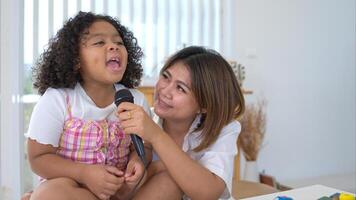 A tiny girl and mother sits on a toy table, clutching a microphone, and sings cheerfully in the living room photo