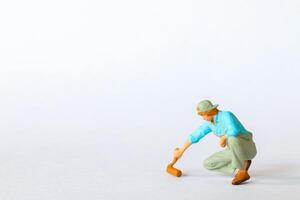 Miniature figure of a painter in a blue shirt, green overalls, crouched while using a roller brush. on a white background photo