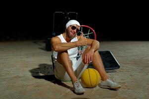 A basketball player sits on the court wearing sunglasses, relaxed after an intense practice session at dusk photo