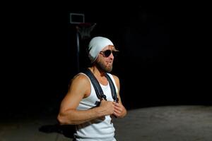 A muscular man wearing sunglasses and a beanie while holding backpack stands confidently in the dark photo