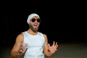 A man wearing a tank top, sunglasses, and a beanie poses confidently at night against a dark background photo